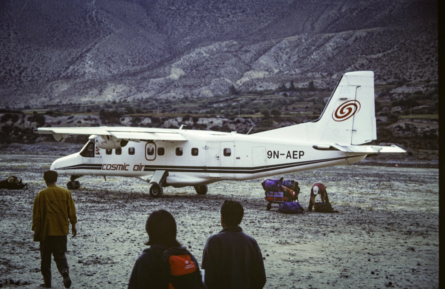 Cosmic Air Twin Otter aircraft 9N-AEP parked at remote Himalayan mountain airstrip with passengers and cargo, showing typical Nepal domestic aviation infrastructure